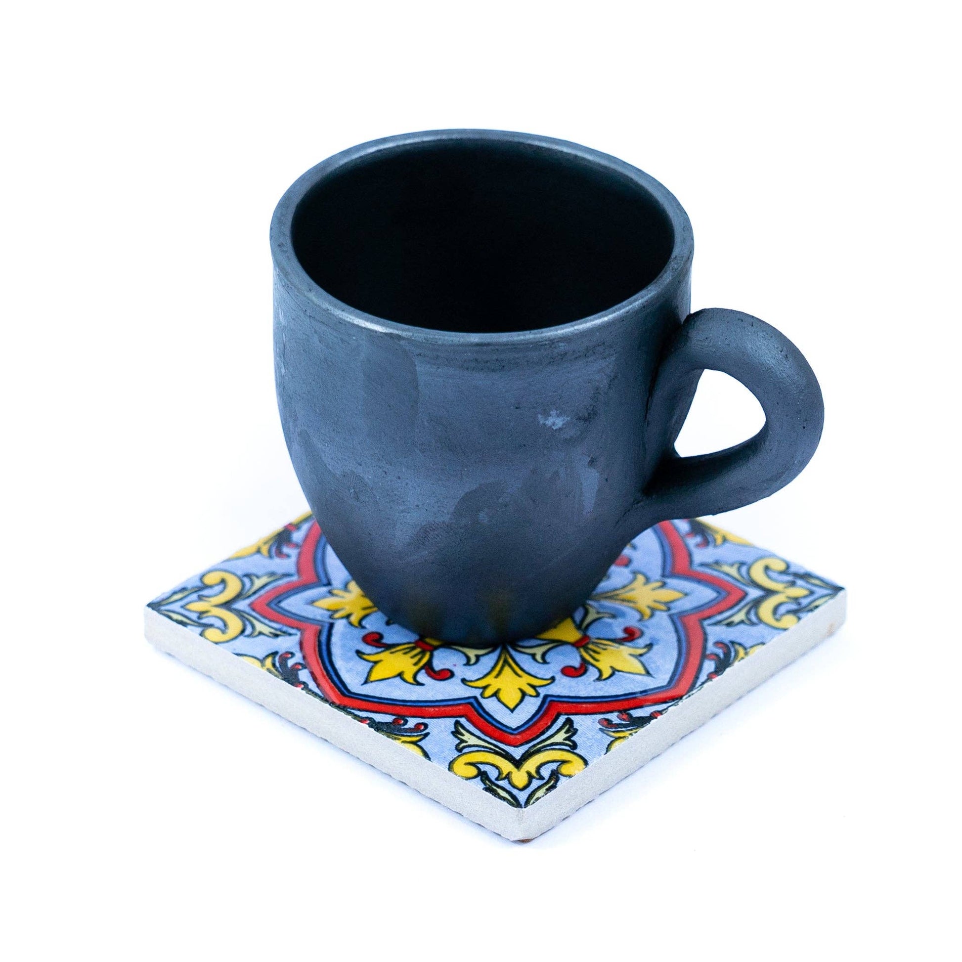 A black ceramic mug rests on a Tile-Inspired Ceramic & Cork Coaster from Meninas Bonitas Cork, featuring a decorative red, yellow, and blue pattern on a square base, set against a white background.