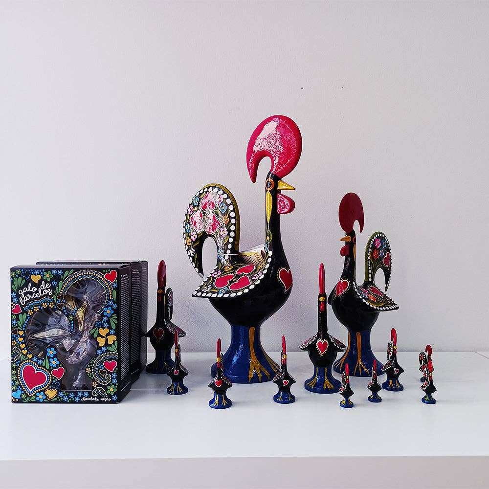 Tejo Shop’s Barcelos Rooster figurines, symbols of Portuguese art, in various sizes with heart and floral patterns, are displayed on a white shelf alongside a decorative box.