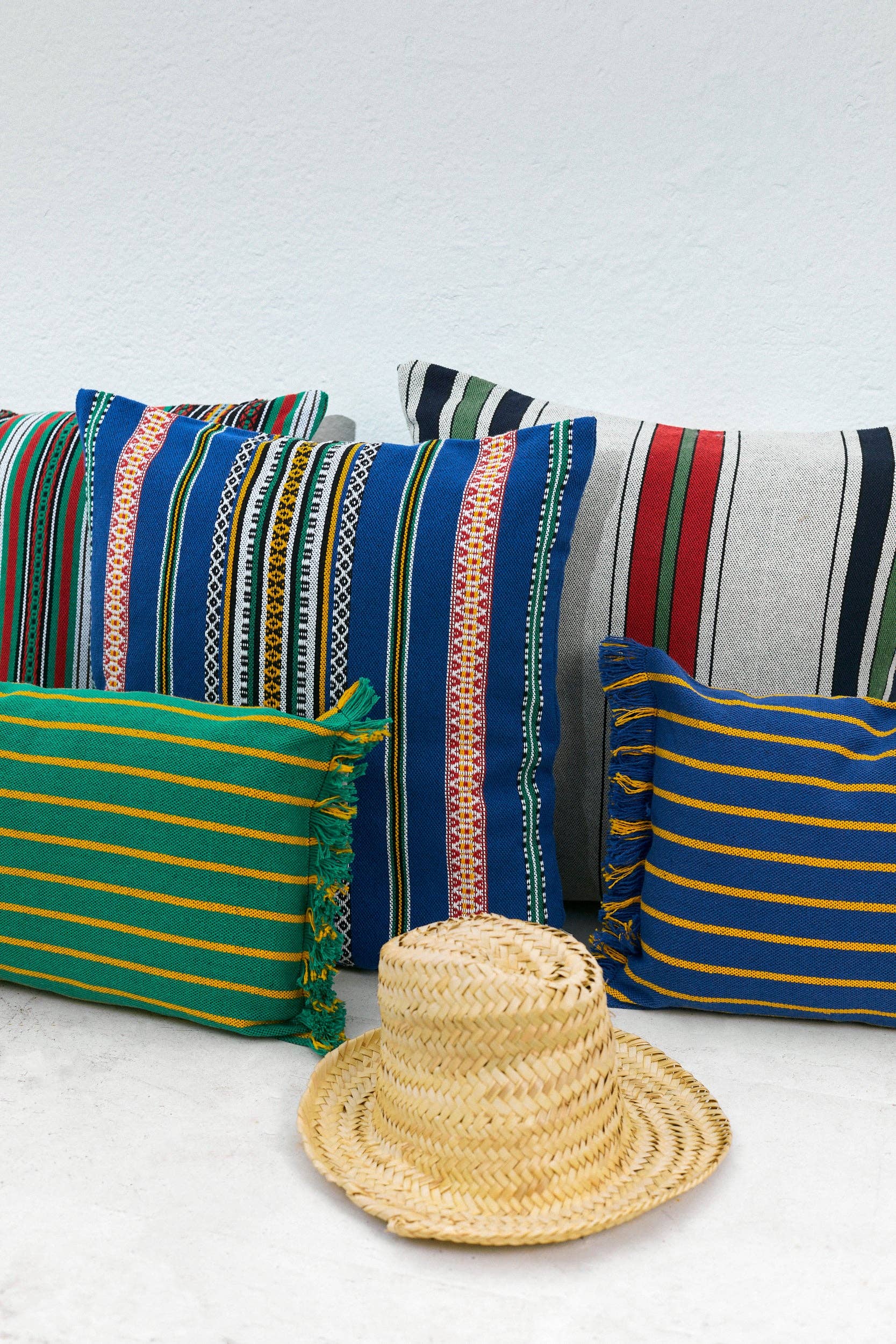 A Blue Pomegranate Cushion Cover (50cmx45cm) by Eturel Madrid is displayed among patterned, Alhambra-inspired pillows against a white background, with a woven straw hat placed in front.