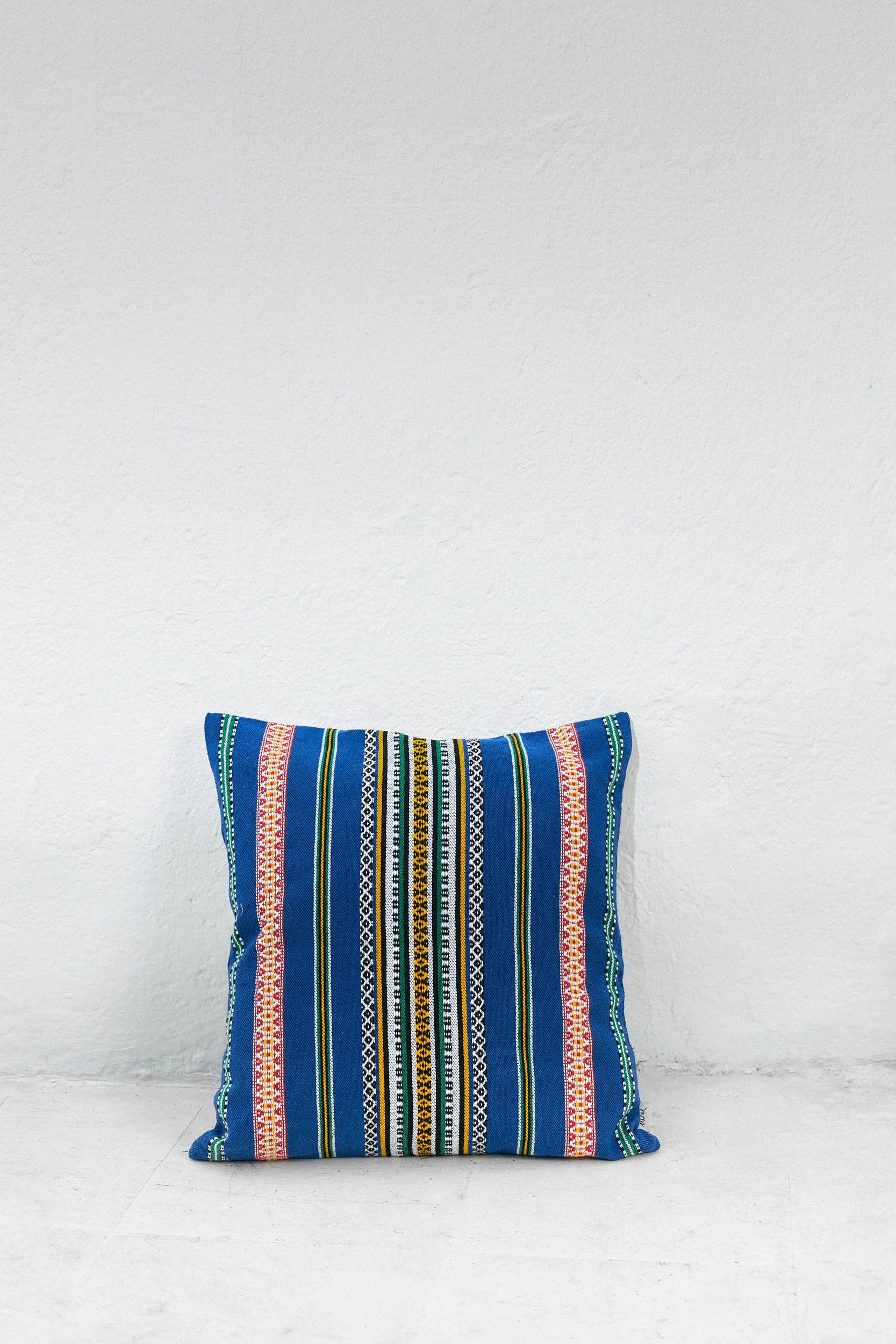 The Eturel Madrid Blue Pomegranate Cushion Cover (50cmx45cm), handmade in Spain and featuring Alhambra-inspired colorful stripes, is displayed against a white textured wall on a light floor.