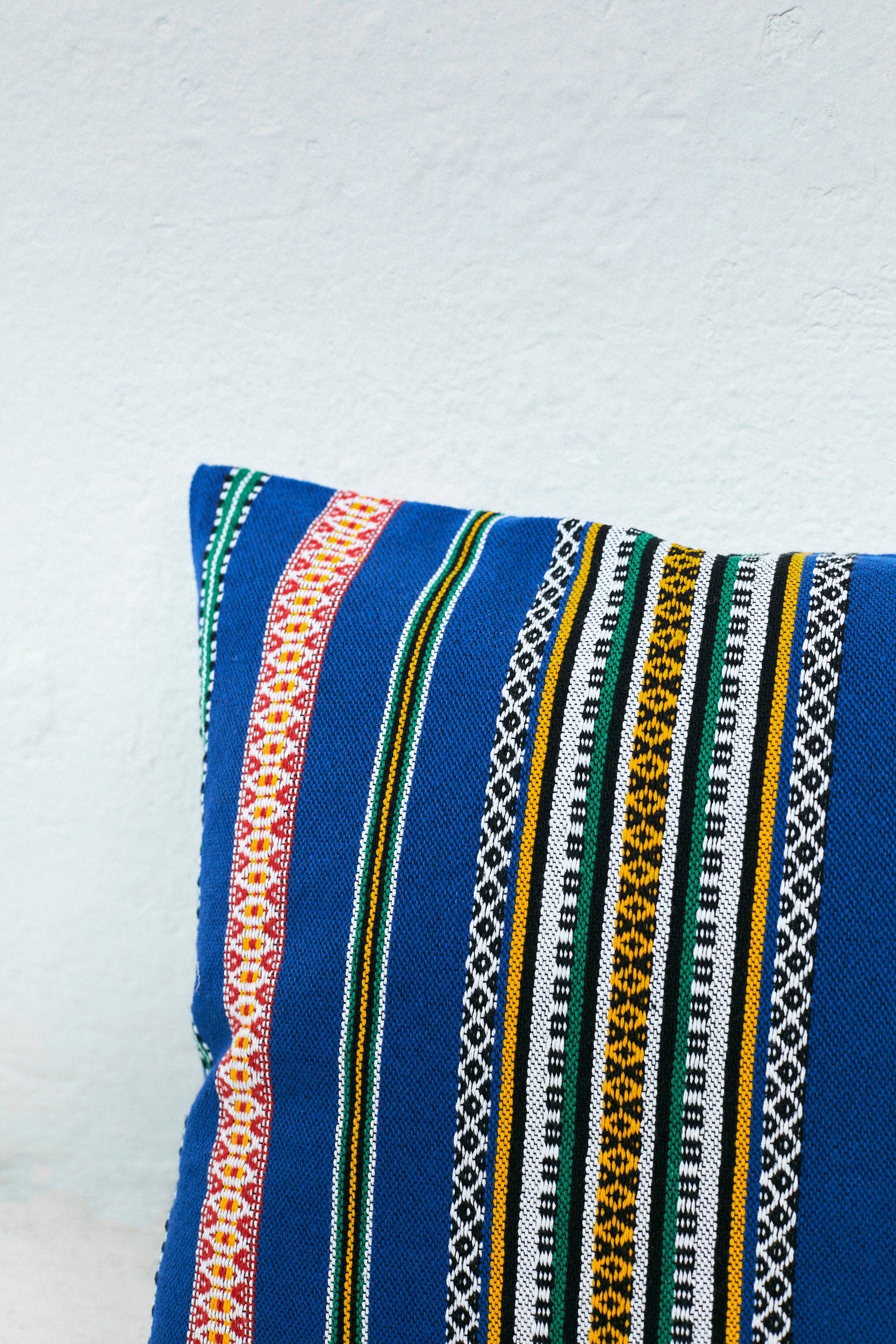The Eturel Madrid Blue Pomegranate Cushion Cover 50cmx45cm, Alhambra-inspired and handmade in Spain, displays colorful geometric stripes and is shown against a light textured wall.