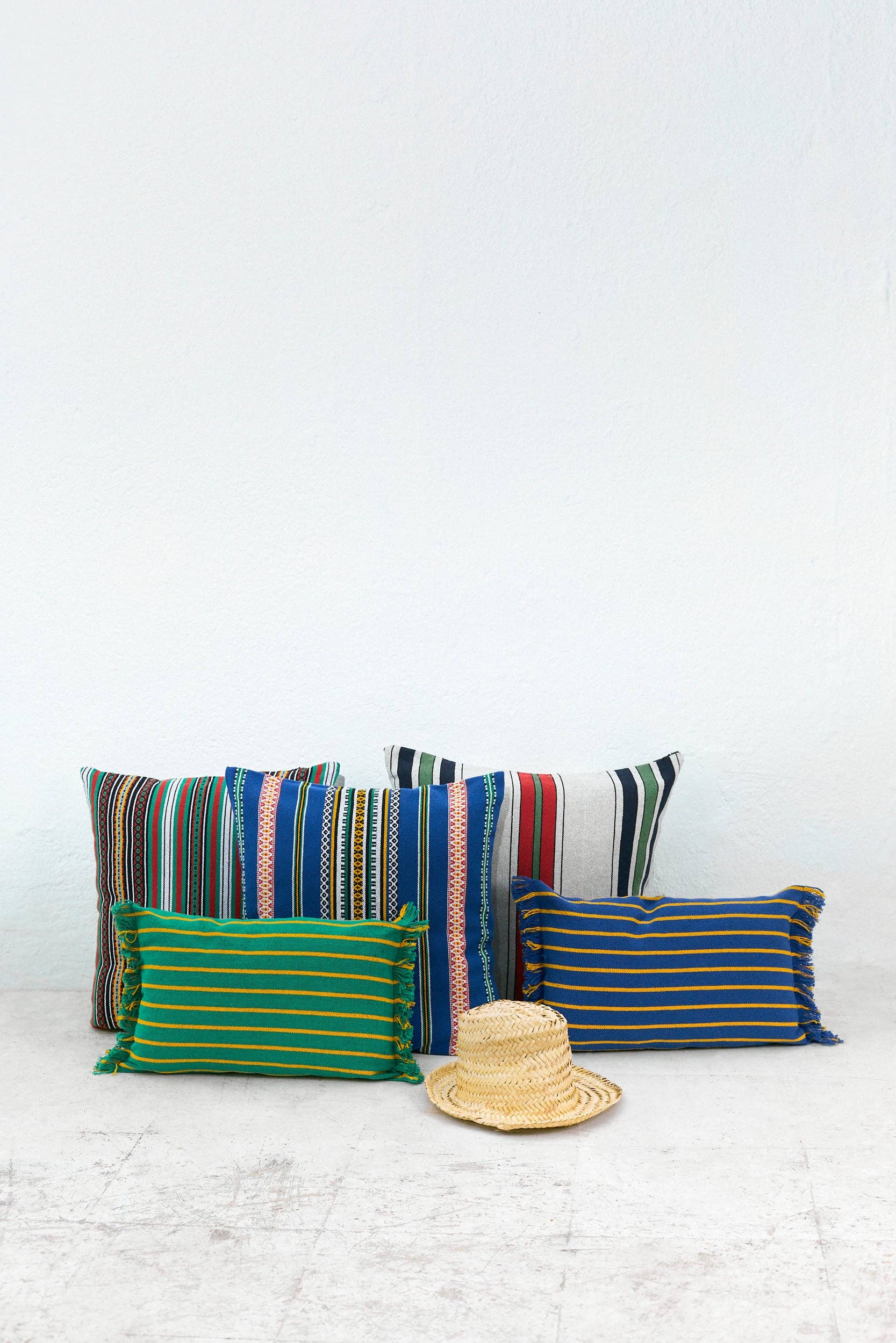 Five vibrant striped pillows, including the Blue Pomegranate Cushion Cover 50cmx45cm by Eturel Madrid, are set against a white wall on a light floor with a woven straw hat in front. Handmade in Spain.