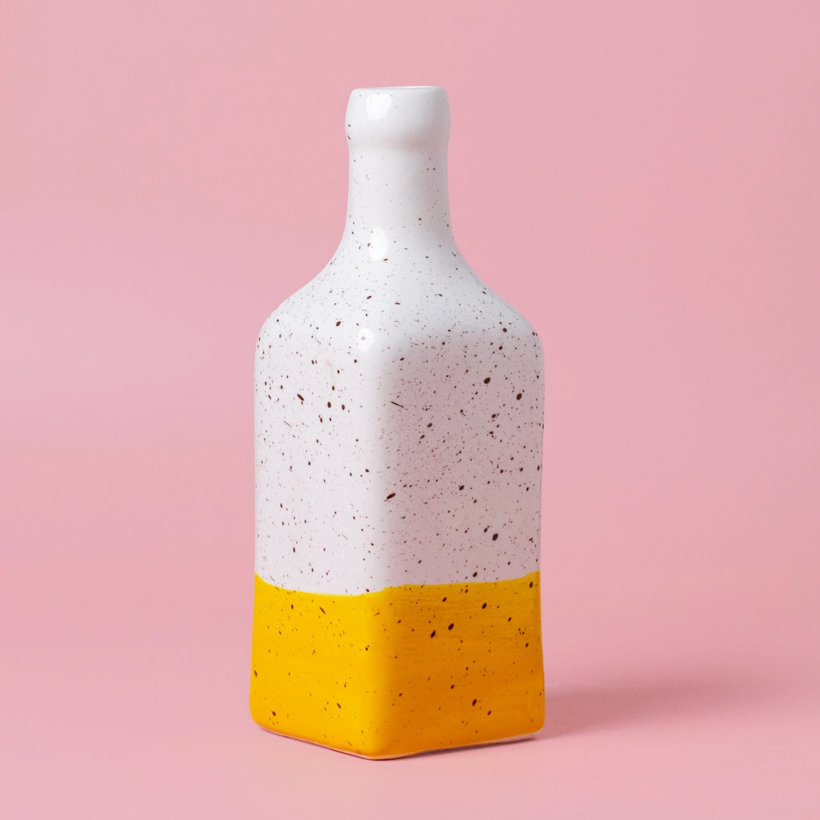 The Salad kit | CANELA Oil and Salad Bowl by Alfar Tierra Cocida features a tall, square ceramic bottle with a speckled white top and solid yellow base, ideal for handmade kitchenware collections, set against a pastel pink background.