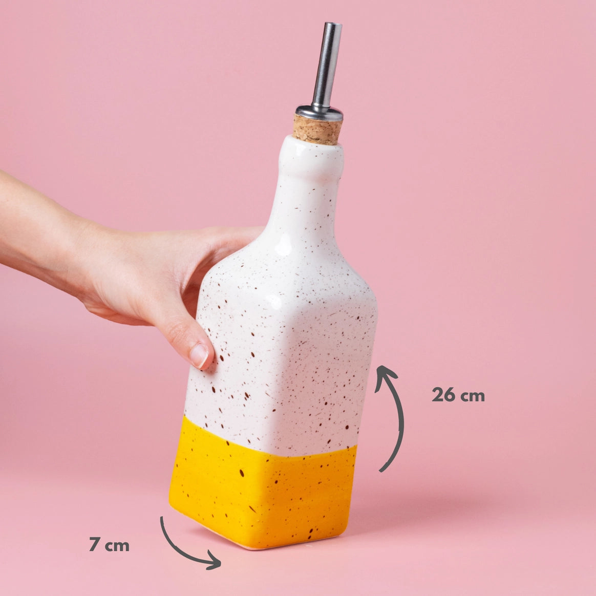 A hand holds the CANELA Oil and Salad Bottle from Alfar Tierra Cocida’s handmade kitchenware collection, speckled white and yellow, 26 cm tall and 7 cm wide, shown against a pink background. Part of the Salad kit.