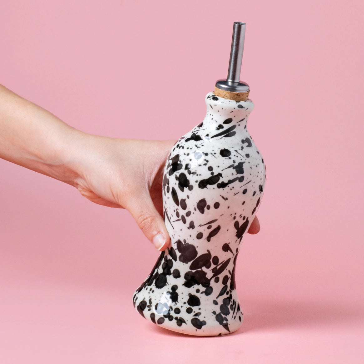 A hand holds the GALAXY Breakfast Grater and Oil Pourer Kit by Alfar Tierra Cocida. The white pourer with black spots and a metal spout stands out against a pink background—a perfect kitchen gift for any home chef.