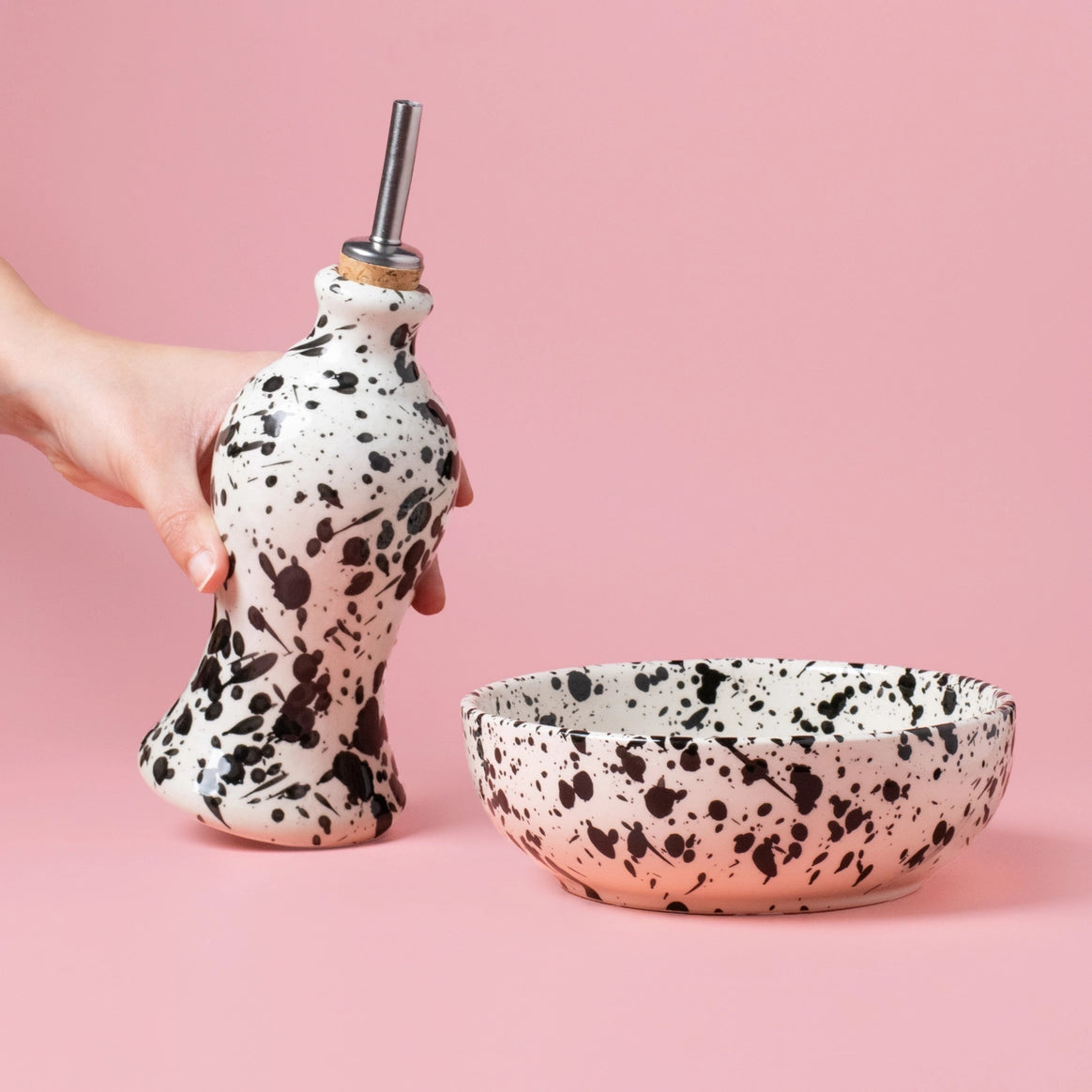 A hand holds Alfar Tierra Cocidas GALAXY Breakfast Grater and Oil Pourer Kit, featuring speckled ceramic pieces beside a matching bowl—an ideal kitchen gift—set against a pink background.