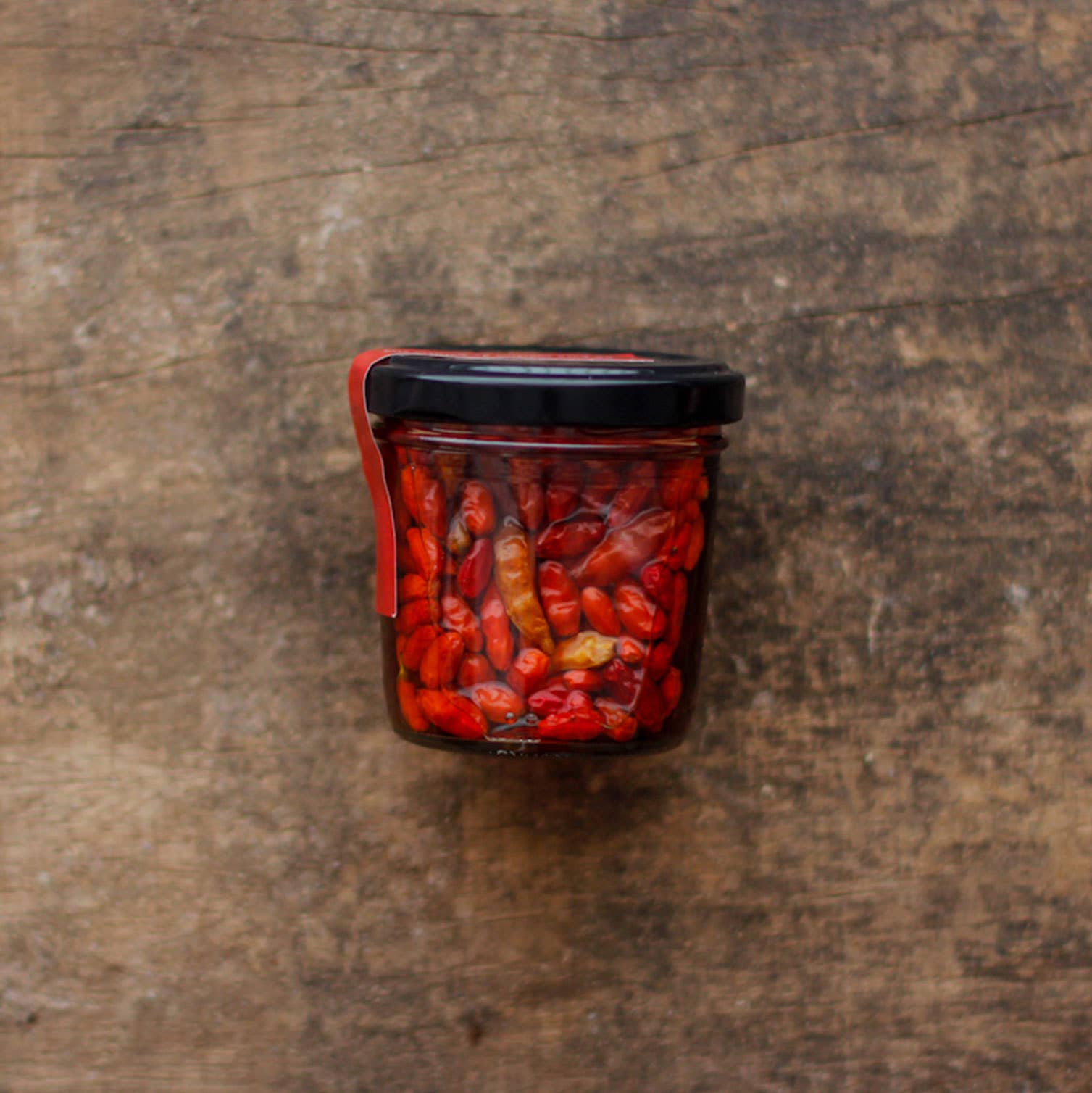 The Lisbon Essentials Box by Iberica - Pretty things from Portugal features a small glass jar of red chili peppers with a black lid, ideal for making Piri Piri oil, displayed on a rustic wooden surface.