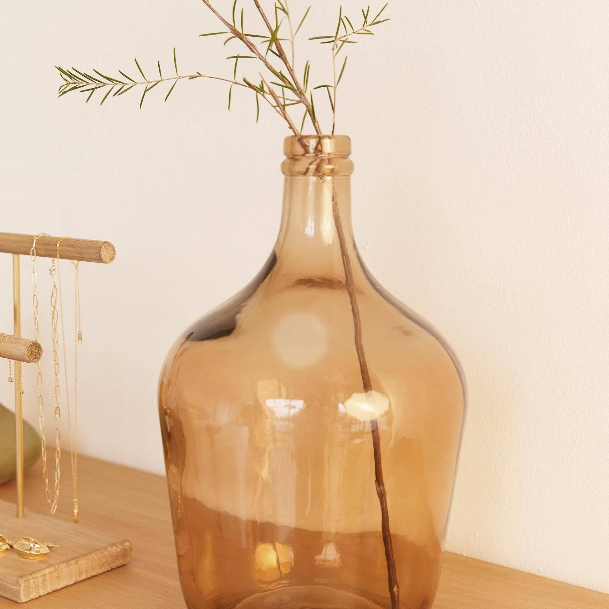 The Hannun Luiza Demijohn Vase, large and amber-colored with a narrow neck, sits on wood holding a thin branch with sparse leaves. Nearby, a wooden jewelry stand displays delicate necklaces.