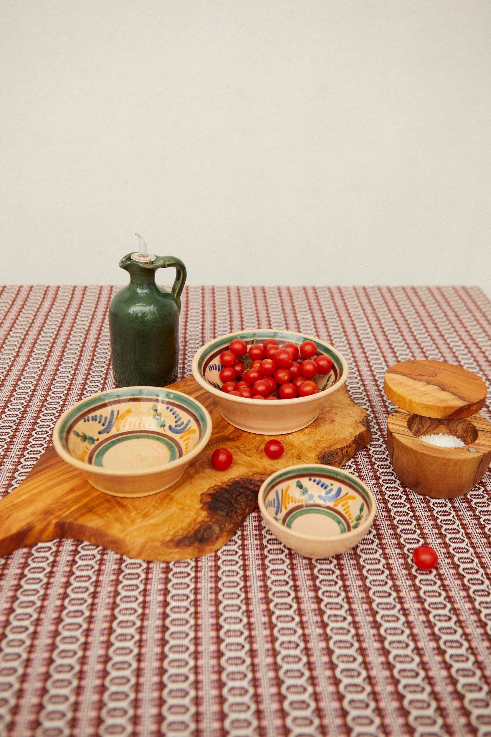 A jug of olive oil, a bowl of cherry tomatoes, an Eturel Madrid Medina Printed Ceramic Bowl, and a wooden salt container sit on a wooden board atop a red and white patterned tablecloth, reflecting Córdoba heritage.