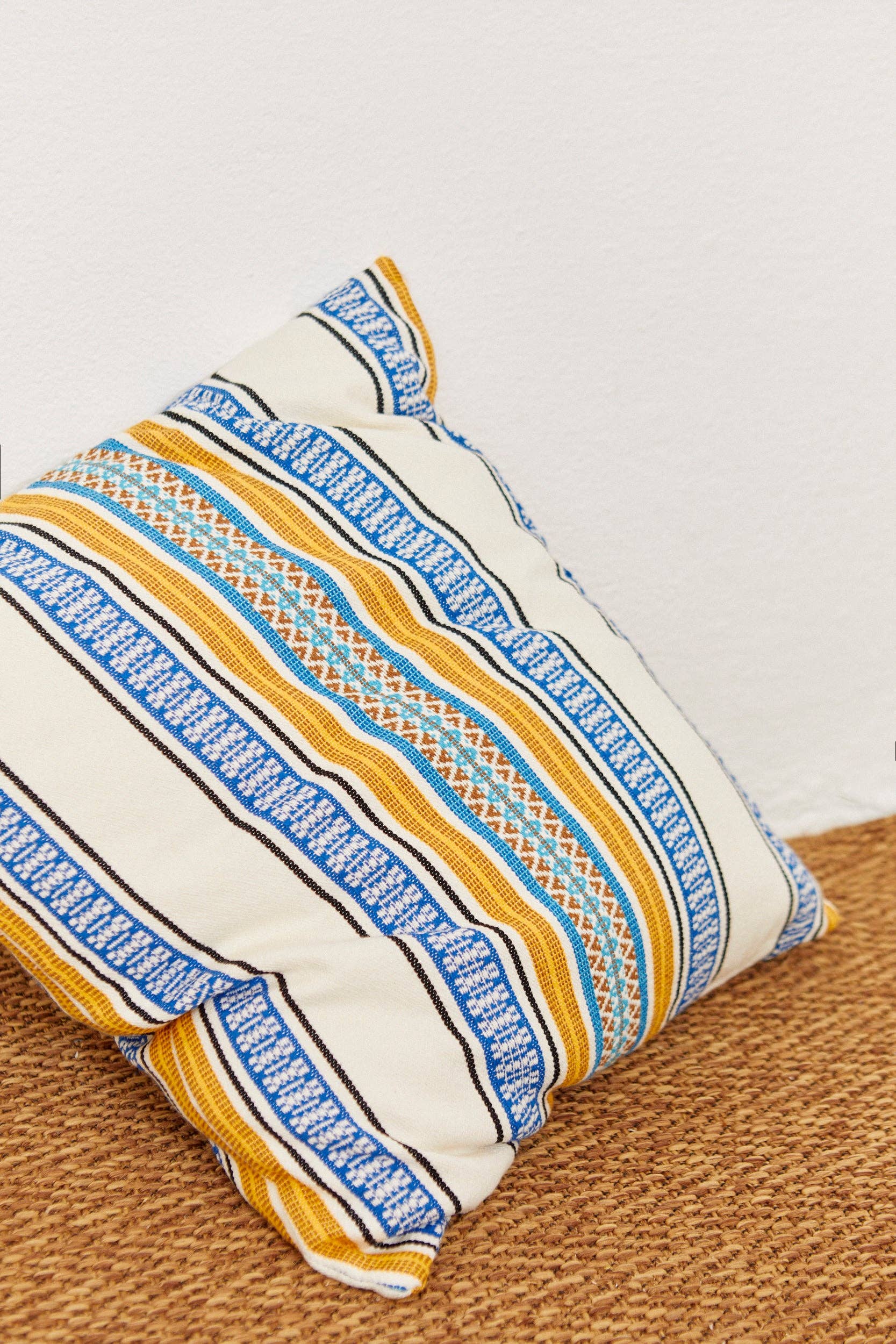 The Eturel Madrid Morellano Blue Woven Cushion Cover 50cmx50cm, featuring blue, yellow, and orange geometric stripes, rests against a white wall on a woven brown carpet.