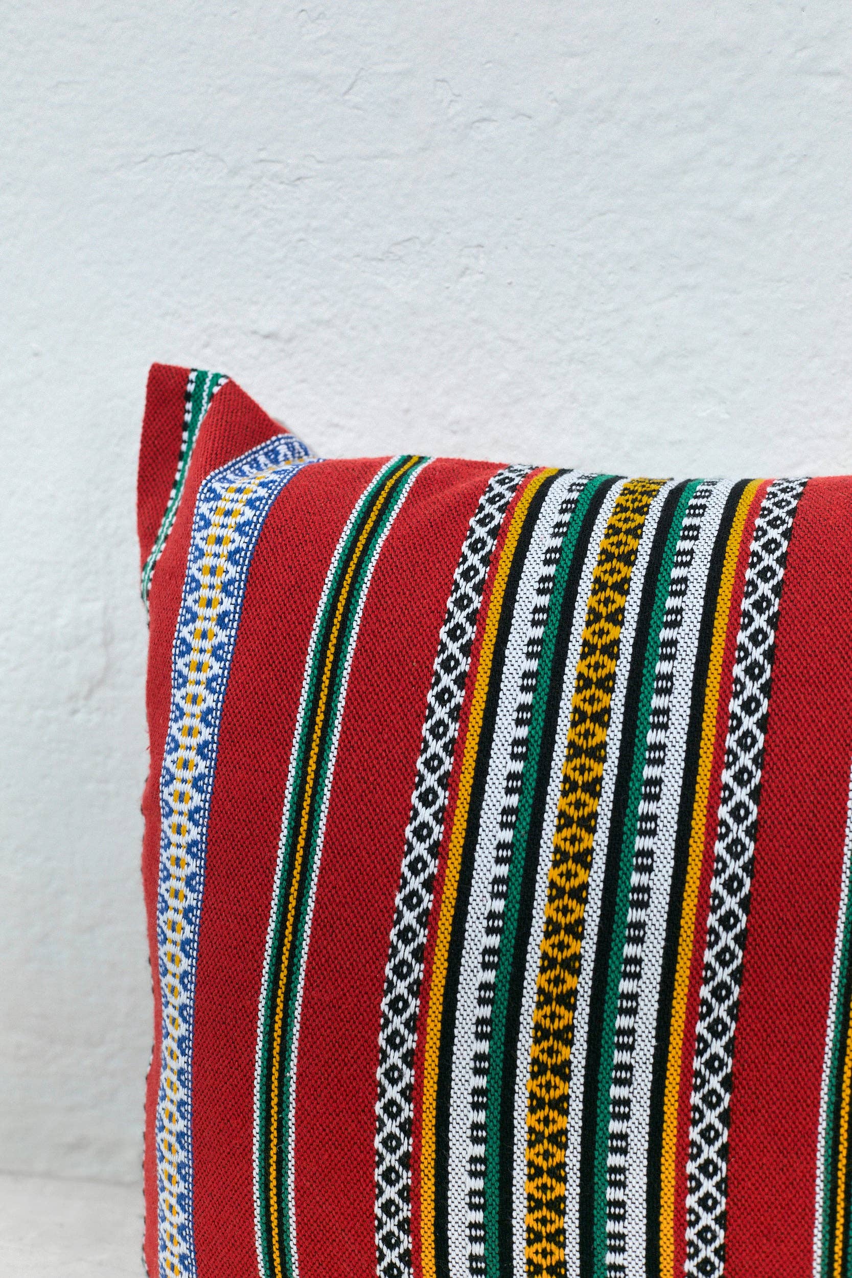 The Eturel Madrid Red Pomegranate Cushion Cover (50cmx45cm) features colorful woven stripes in black, white, yellow, green, and blue, resting against a white textured wall and highlighting Spanish craftsmanship.