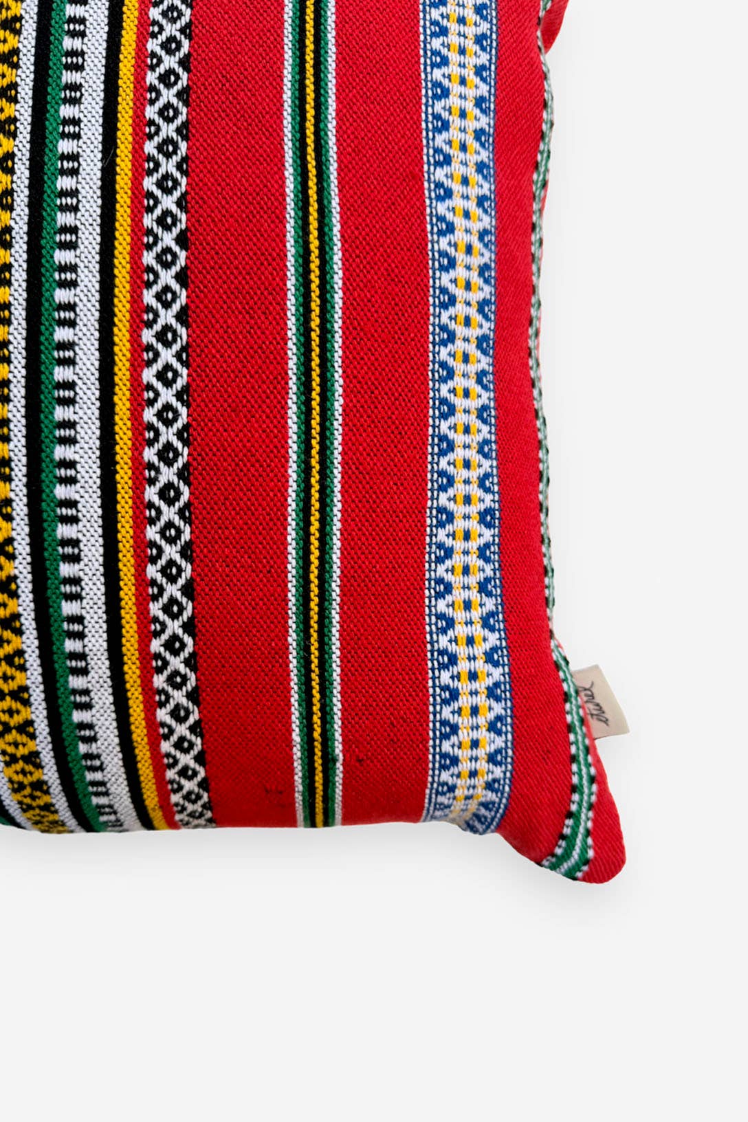 A close-up of the Red Pomegranate Cushion Cover 50cmx45cm by Eturel Madrid, handmade in Spain with vibrant red fabric, decorative woven stripes in black, yellow, green, blue, and white, and a small side fabric tag.