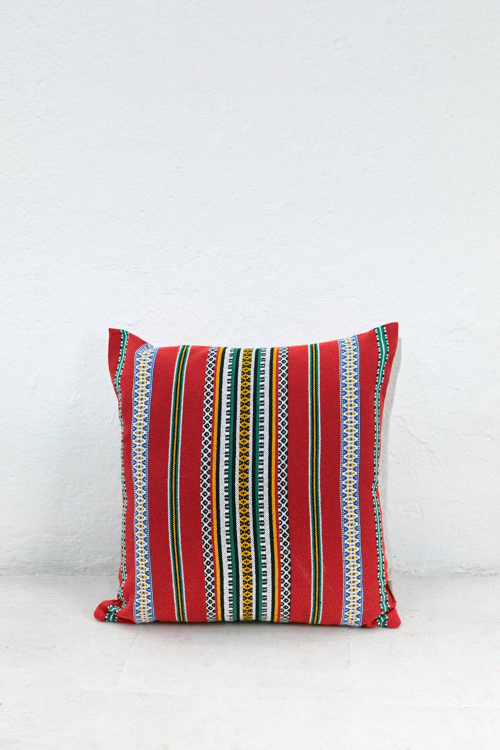 The Eturel Madrid Red Pomegranate Cushion Cover (50cmx45cm) features a vibrant striped design in red, yellow, green, blue, black, and white—highlighting exquisite Spanish craftsmanship against a plain white wall on a light floor.