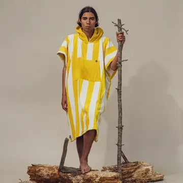 A person stands barefoot on logs, wearing the Futah Bedu Terry Poncho—a yellow and white striped Portuguese cotton poncho with a hood—and holds a tall branch upright against a plain beige background.