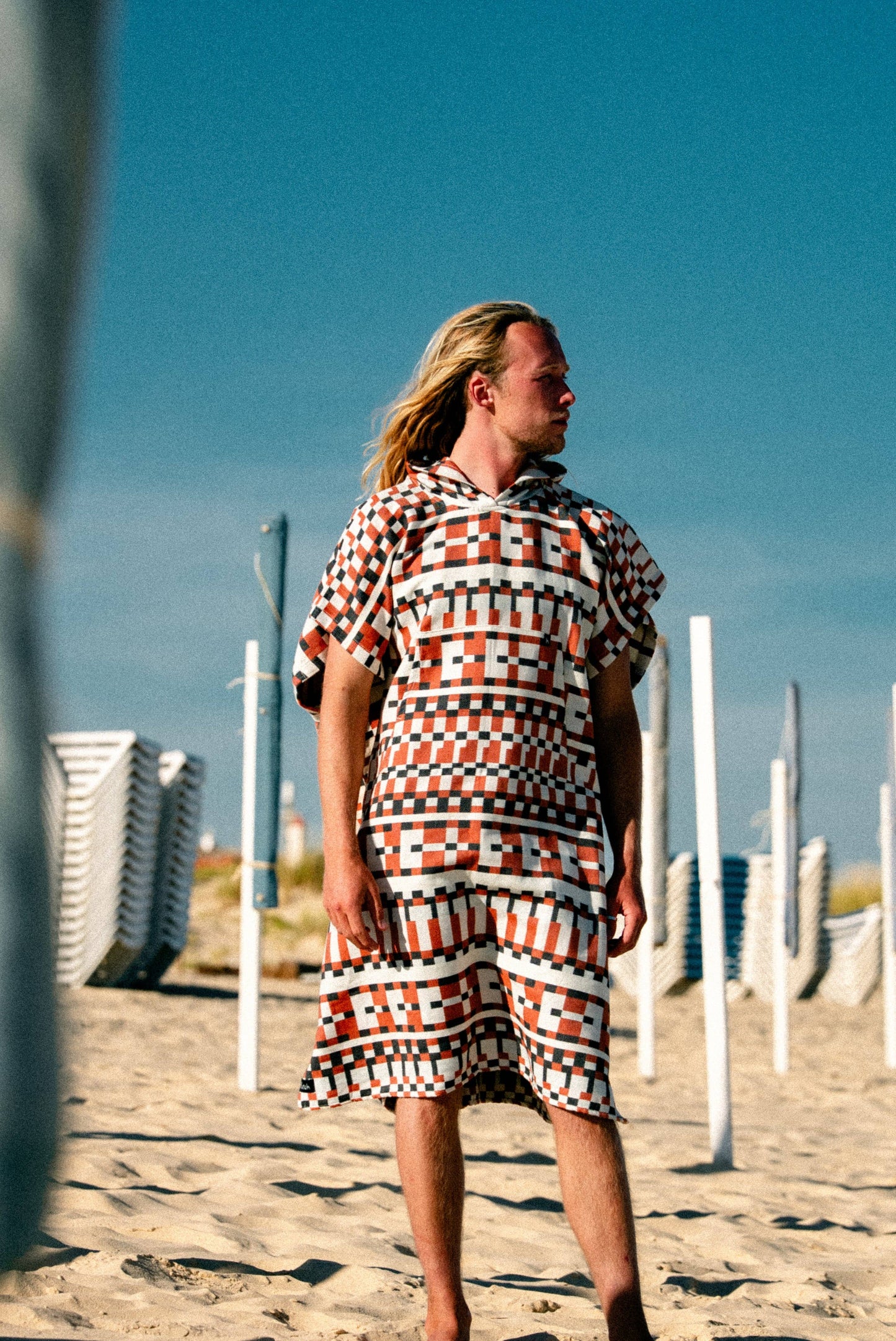 A man with long blond hair stands barefoot on a sandy beach, wearing the Cacela Poncho by Futah—a patterned Portuguese beachwear piece featuring red, white, and black squares. Stacked chairs and poles appear in the background beneath a clear blue sky.
