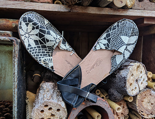 A pair of Valentims Ladies Espadrille Sandals by Iberica, featuring a black and white floral pattern, made in Portugal, rest atop stacked logs and wooden tubes outdoors.