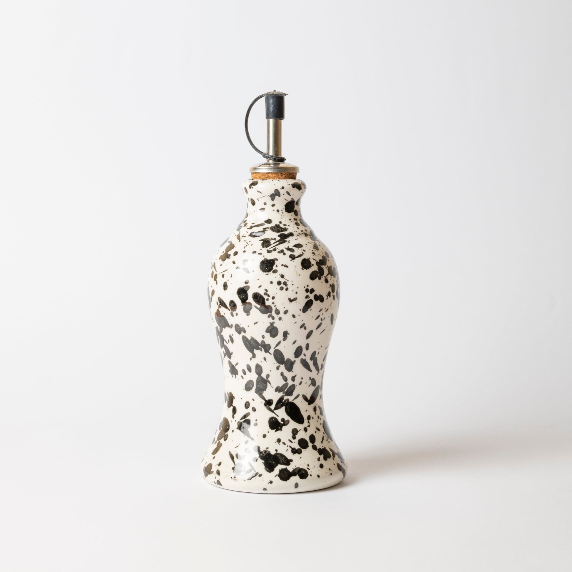 The Galaxy Olive Oil Pourer by Alfar Tierra Cocida features a ceramic white base with a unique black splatter design and a metal spout, creating a modern kitchen accessory. Shown here against a plain white background.