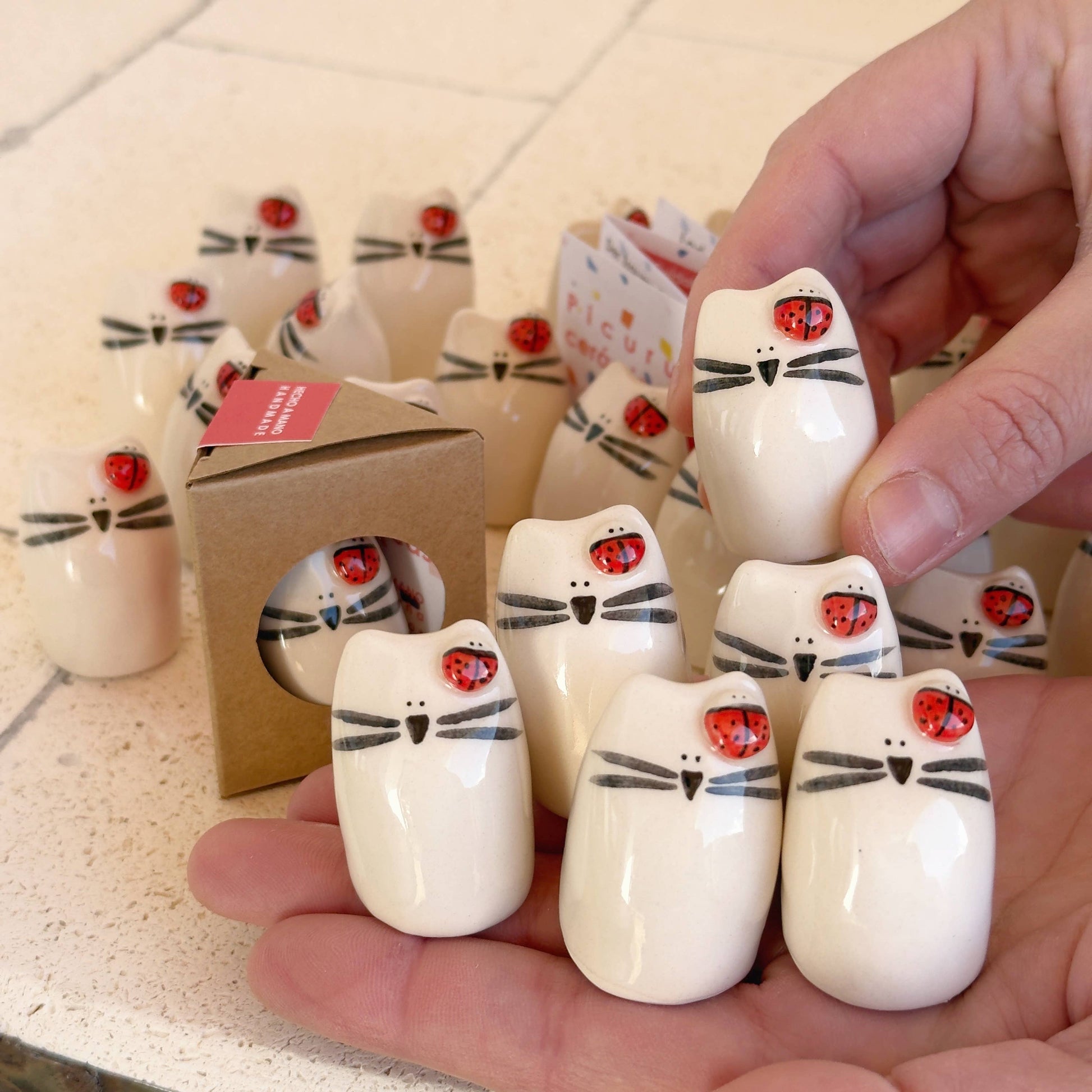 A hand holds several Picuru ceramicas Ladybug Amulet, Guardian of Your Dreams figurines—handmade lucky charms with painted whiskers and tiny ladybugs. More amulets and a small box rest on a light surface in the background.