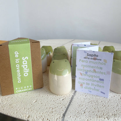 A set of Little Frog Amulet of Adventures by Picuru ceramicas, handmade with green-topped smiling faces, is displayed beside its brown box—making a unique gift and charming symbol of joy and new experiences.