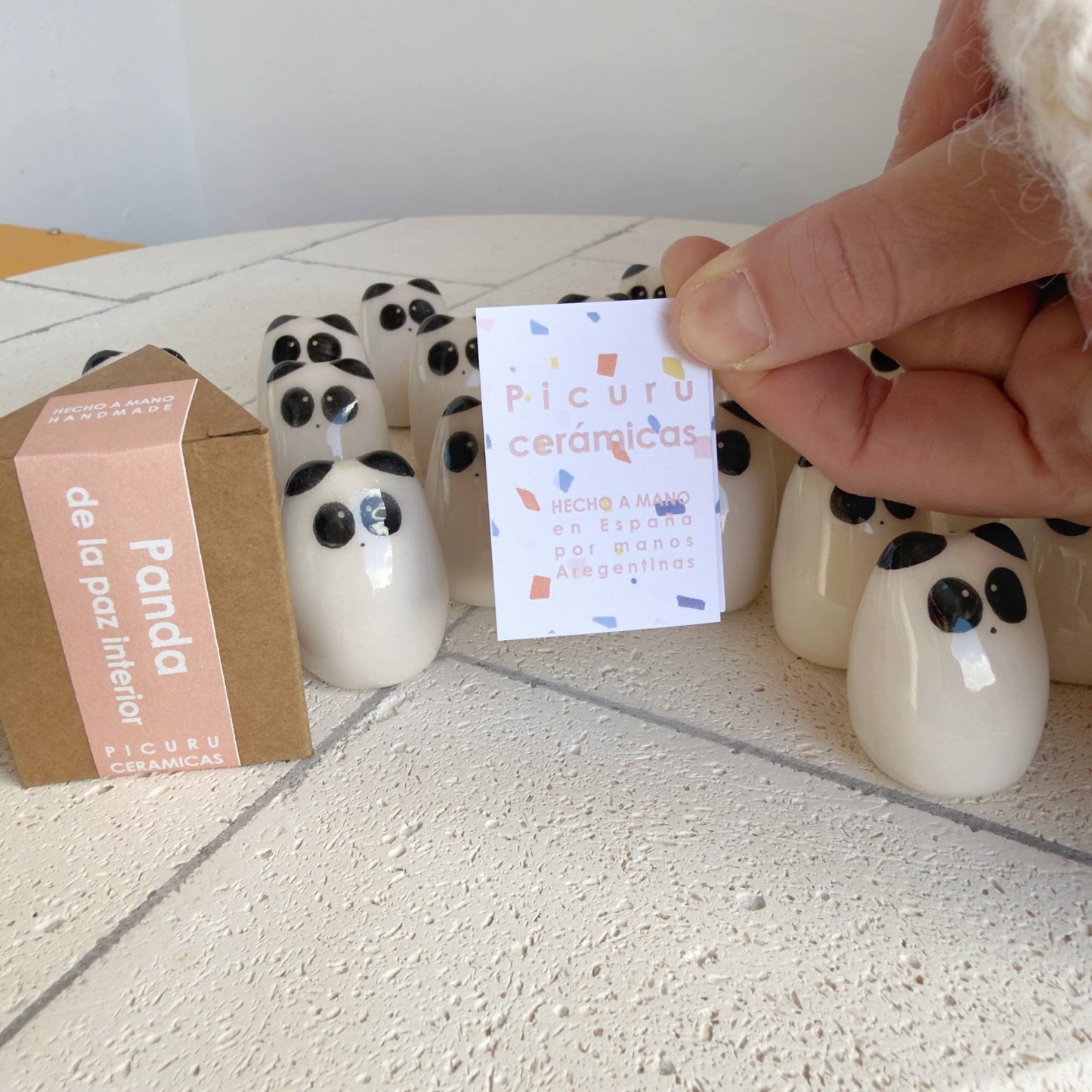 A hand displays a Panda Amulet of Inner Peace – Handmade Lucky Charm Gift by Picuru ceramicas, alongside a branded card and box. These unique items sit on a white textured surface—perfect for gifting tranquility and luck.