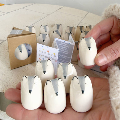 A hand holds several Picuru ceramicas’ Little Wolf Amulet of Courage—handmade white ceramic figures with black beak-like markings, each a symbol of strength and growth. More amulets and their packaging are on the white table in the background.