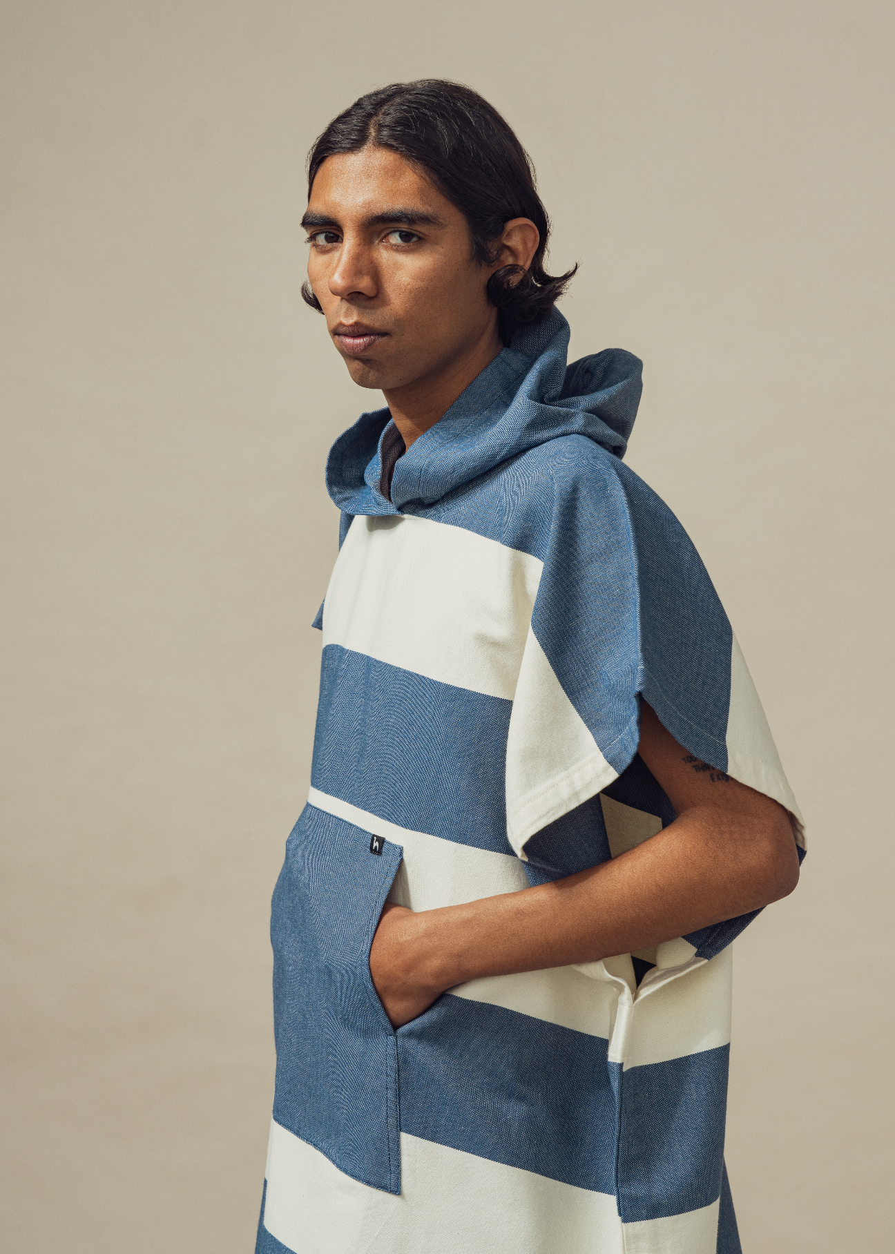 A person with medium-length dark hair wears the Futah Bedu Cotton Beach Poncho – a premium Portuguese surf and beach poncho featuring blue and white stripes and a hood – standing against a plain beige background with hands in pockets, looking at the camera.