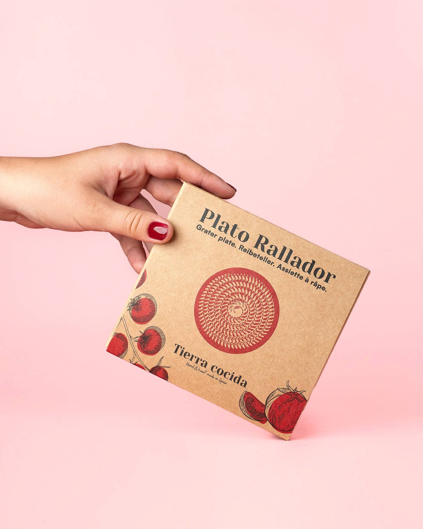 A hand with red nail polish holds a square box labeled Alfar Tierra Cocida featuring the Handmade Ceramic Grater Plate Small - Alcazaba, decorated with tomato illustrations and Spanish text—a unique kitchen gift for grating garlic.