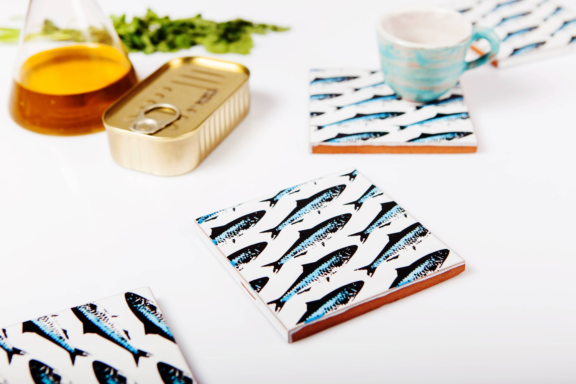 A Bussoga Sardines Coaster with a fish pattern sits on a white surface near a blue cup, an open tin of fish, an oil bottle, and herbs—ideal for anyone who loves unique handmade coasters.