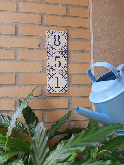 The Personalized Portuguese Tile House Number by TilePassion features handpainted ceramic tiles displaying 8, 5, and 1 on a yellow brick wall, with a blue watering can and leafy plant in the foreground for added charm.