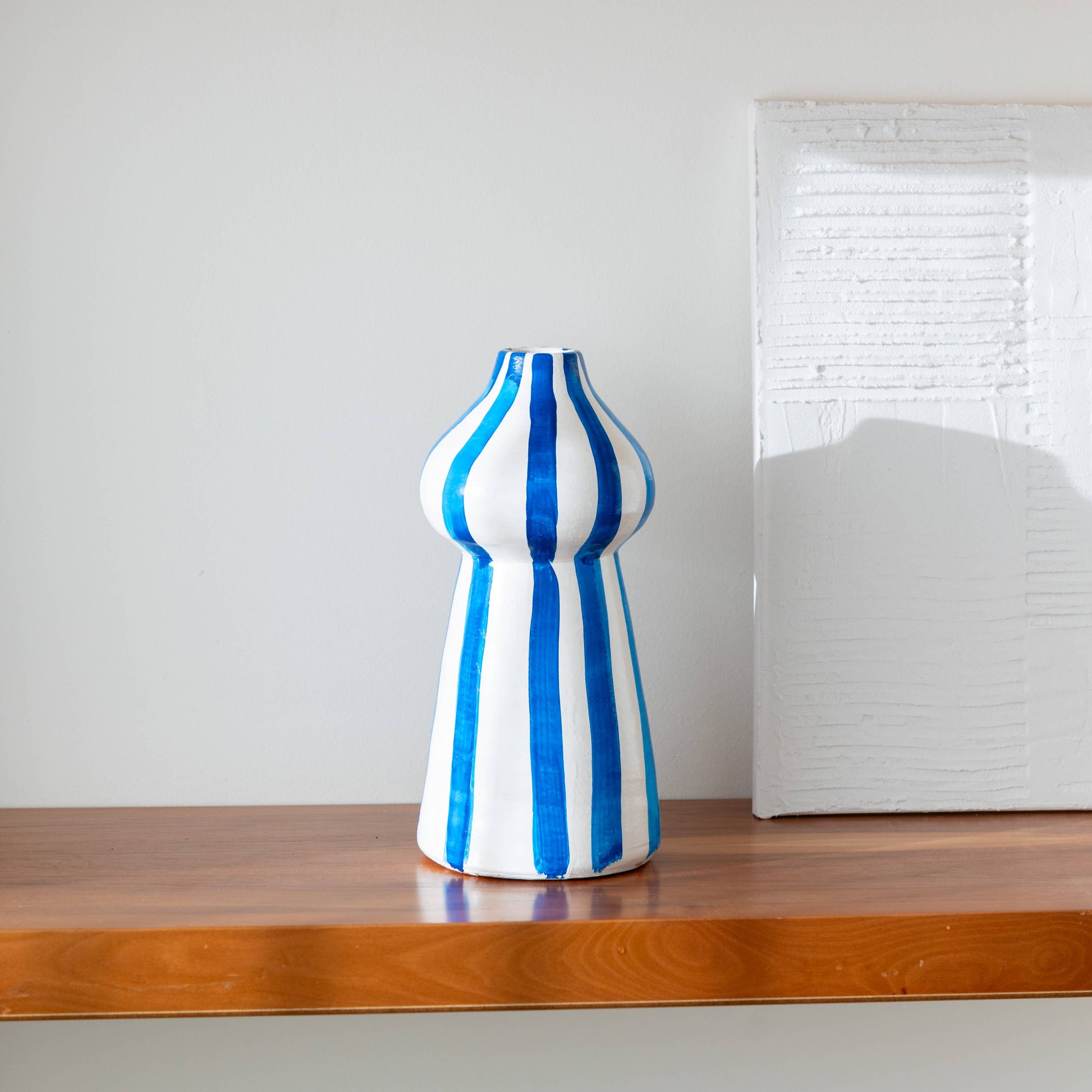 The Lucca vase by OITO Atelier, a handcrafted ceramic piece with blue vertical stripes, rests on a wooden shelf beside a white textured rectangular artwork against a light-colored wall.