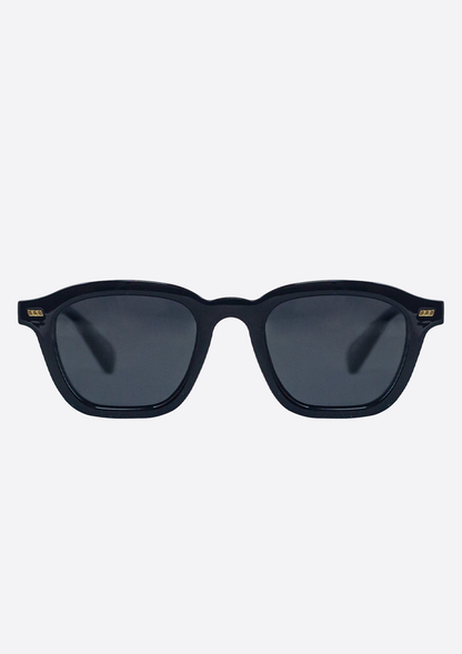 The Oceanides Neda Shiny Black Sunglasses feature a recycled plastic frame, dark polarized lenses, and gold corner accents, displayed against a plain light background.