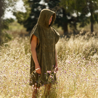A person wearing the Futah Ericeira Terry Poncho, a sleeveless olive-green cotton hooded piece, stands in a sunlit field of tall grass and wildflowers with blurred trees in the background.