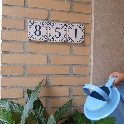 A person waters green plants with a blue watering can near a brick wall displaying house number 851 on a TilePassion Personalized Portuguese Tile House Number – Handpainted Custom Address Sign.