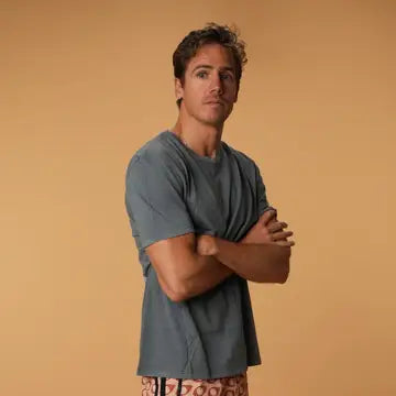 A man with short brown hair stands against a tan background, confidently crossing his arms while wearing the Praia T-Shirt by Futah and patterned shorts, embodying a relaxed, beach-inspired style.