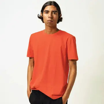 A person with long dark hair wears the Futah Praia T-Shirt in orange, paired with black pants, standing against a plain light background with hands in their pockets.