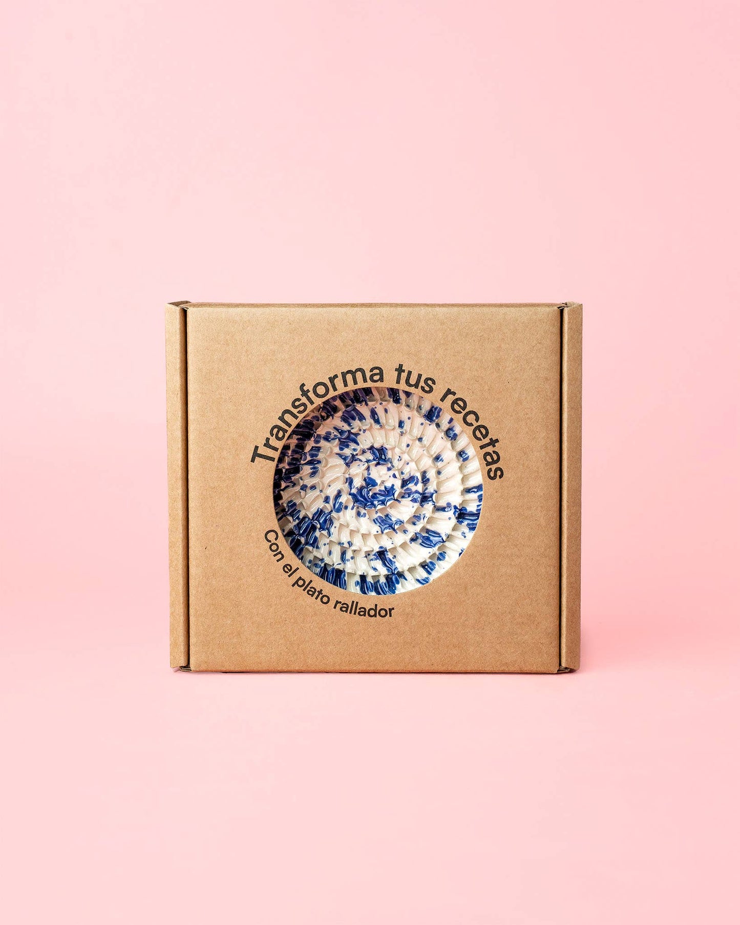 A brown cardboard box with a circular window showcases a blue and white Handmade Ceramic Grater Plate Small - Alcazaba by Alfar Tierra Cocida. Spanish text on pink reads: Transforma tus recetas con el plato rallador—an ideal kitchen gift.