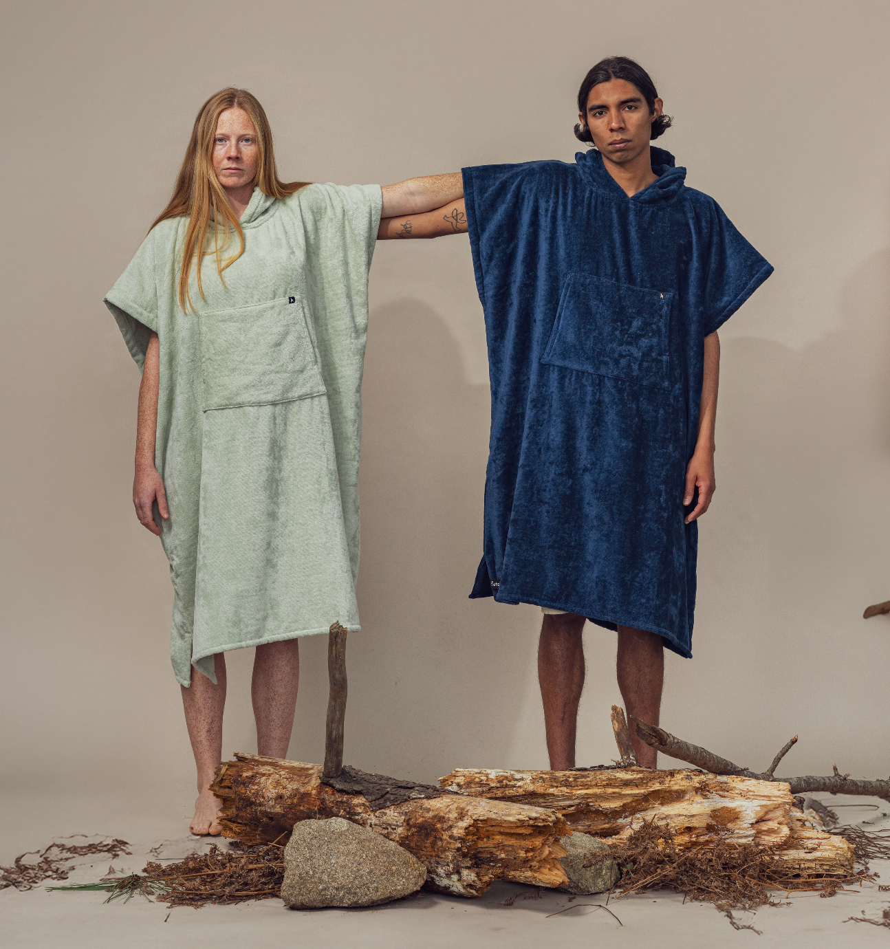 Two people wear Futah's Ericeira Terry Poncho—one light green, one dark blue—standing side by side with arms outstretched among logs and branches on a neutral background. Made in Portugal.