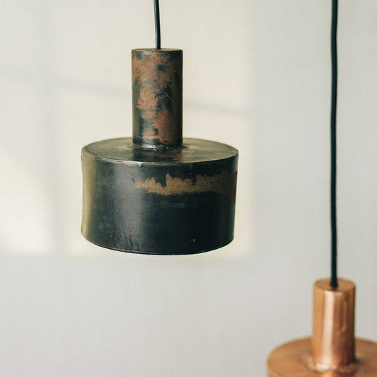 A close-up of two modern Seber Lamps by Hannun, featuring a rustic industrial design in hand-forged iron. Shown here in weathered black and copper tones, these pendant lights hang on black cords and are available in three colours.