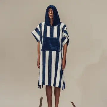 A person stands barefoot, wearing the Futah Bedu Terry Poncho—a quick-drying surf poncho with wide vertical navy blue and white stripes, a spacious front pocket, and a cozy hood—against a plain beige background.