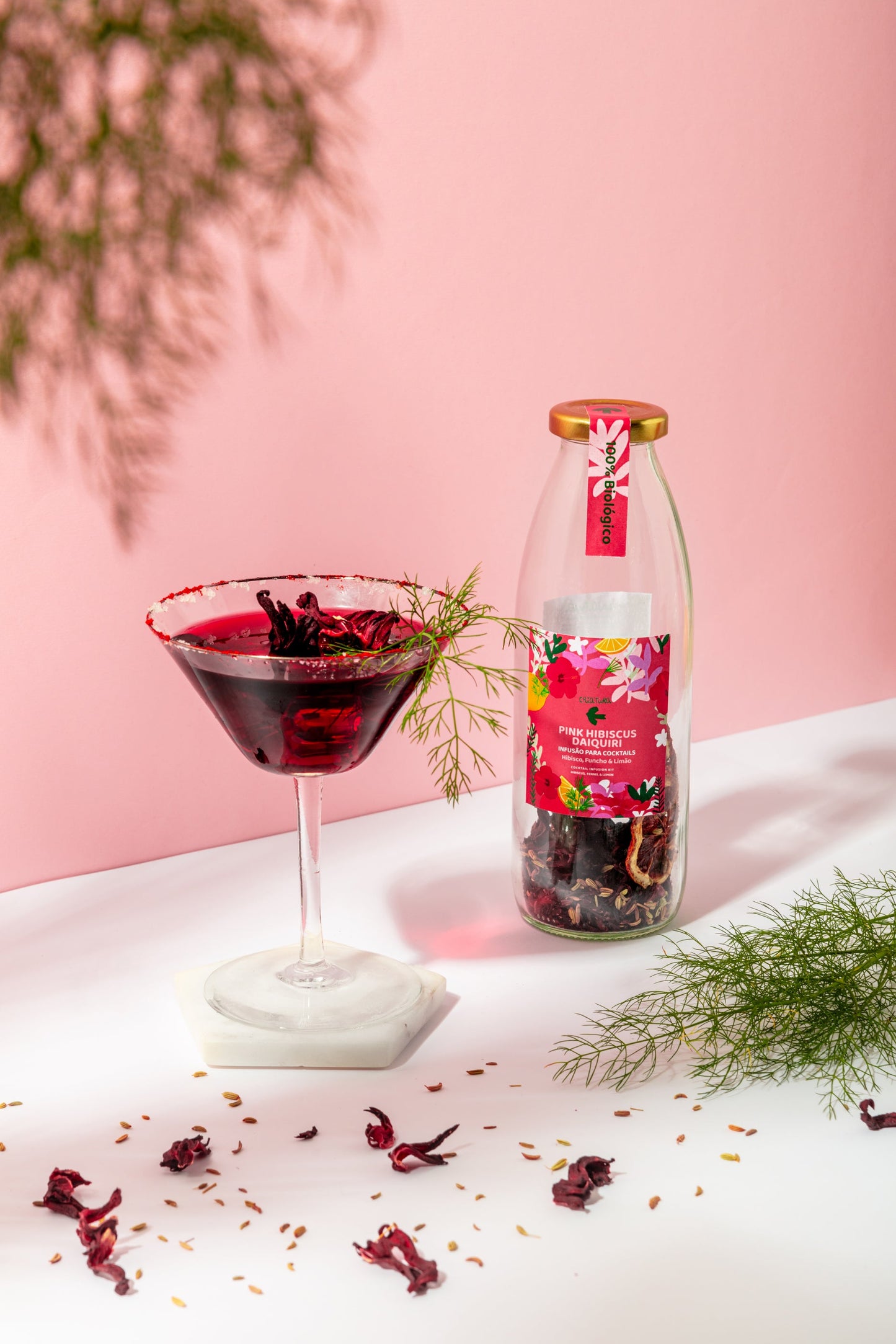 A glass of Pink Hibiscus Daiquiri from Criatura, garnished with herbs, sits beside its bottle on a white surface with scattered petals, set against a pink backdrop and green foliage—ideal for organic cocktail lovers.