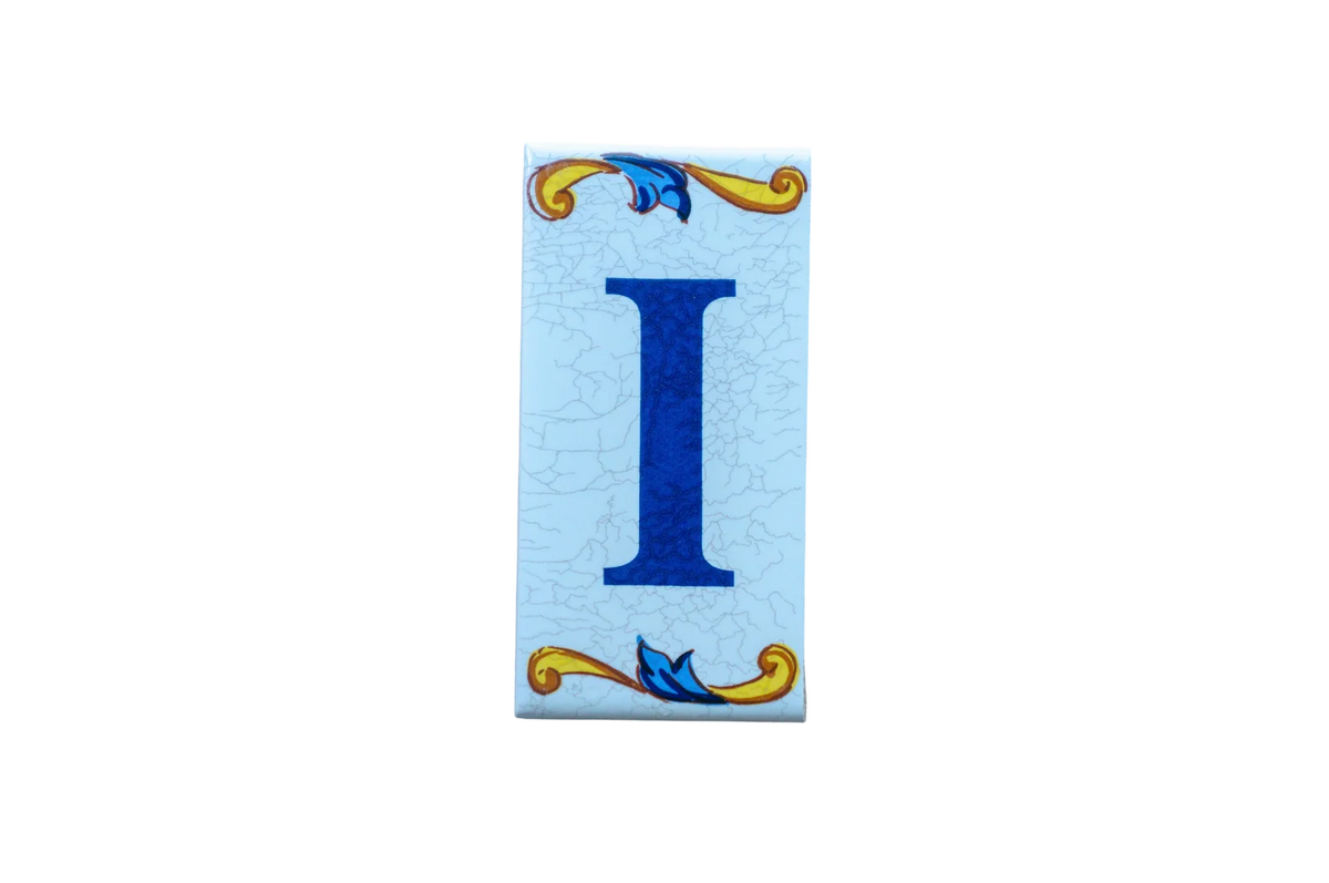 Lisa Croquets Letters – Ceramic House Letter I tile on a light blue crackled background with decorative yellow and blue scrolls at the top and bottom.