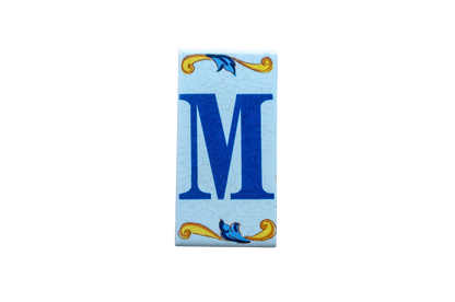 Lisa Croquets Letters – Ceramic House Letter Tile featuring a rectangular Spanish design with a blue letter M and decorative yellow and blue scrolling flourishes at the top and bottom.