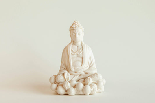 The Manulena Decorative & Aromatic Buddha Piece (White) sits cross-legged on a lotus base with detailed robes and beads, crafted from white ceramic, set against a plain light background.