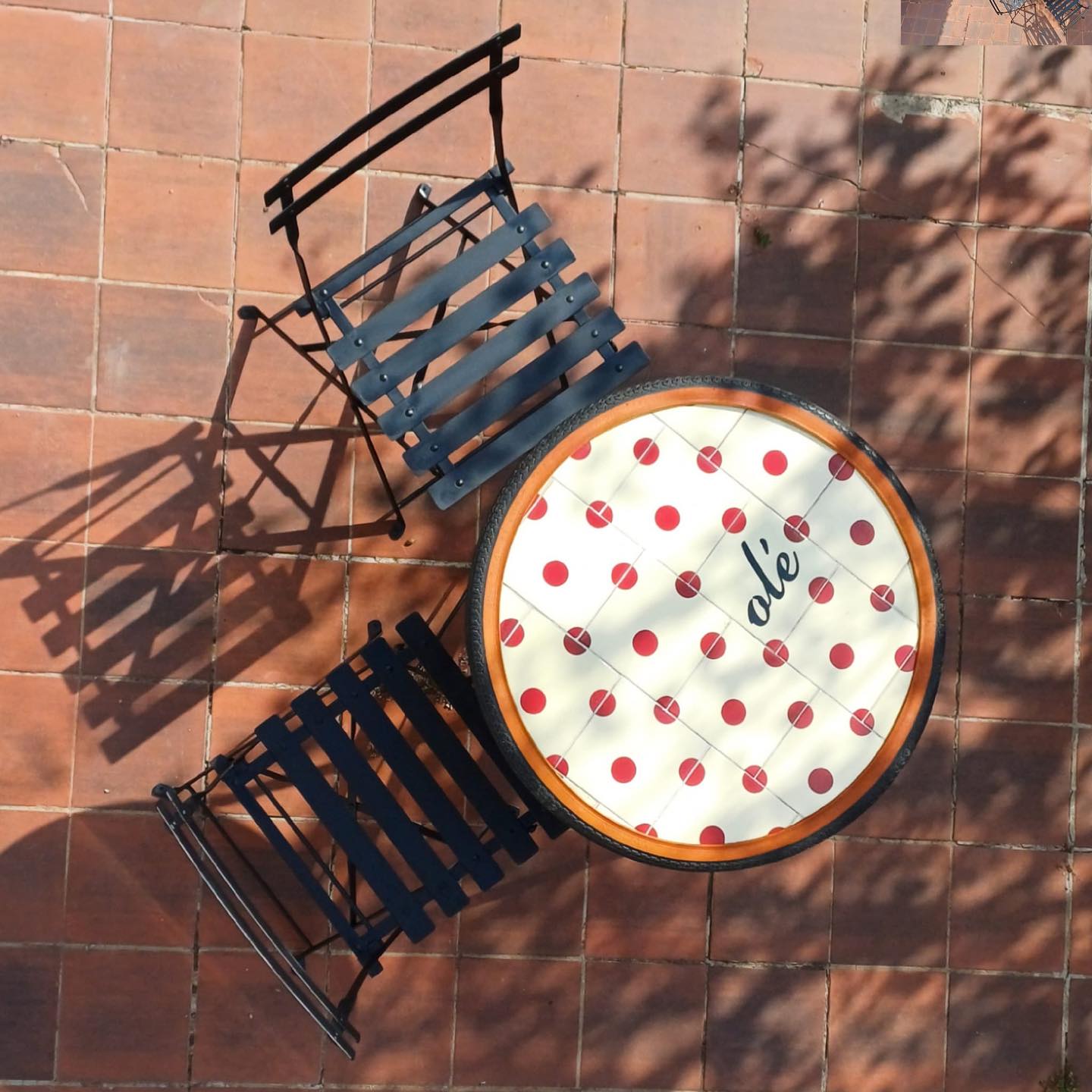Overhead view of two black metal chairs and a round table with a red and white polka dot tablecloth featuring "ole," on a tiled patio. Bussoga's Olé Tile Coasters peek from beneath as tree shadows fall across the scene.