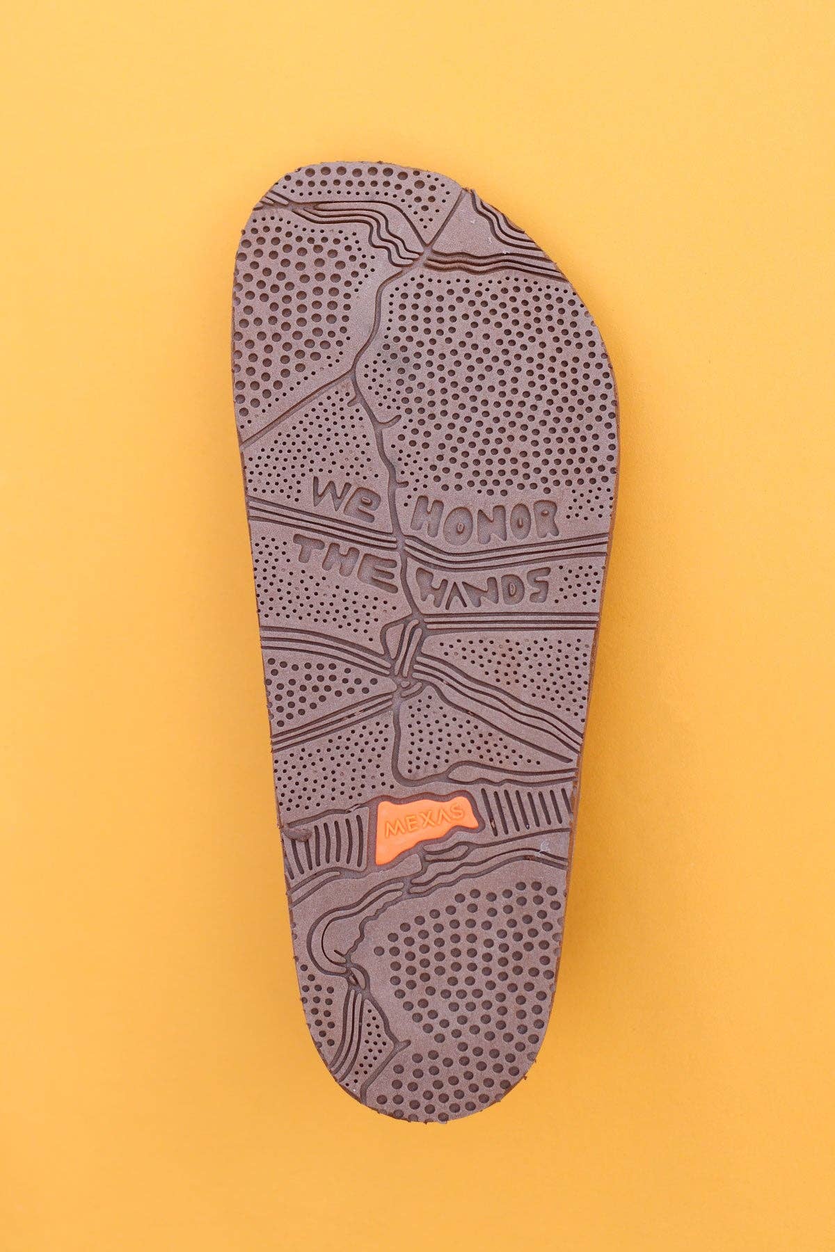 A brown Tampico sandal sole by Mexas EU, featuring textured patterns and "WE HONOR THE HANDS" carved on it, is displayed against a mustard yellow background, highlighting its tribute to handcrafted tradition.