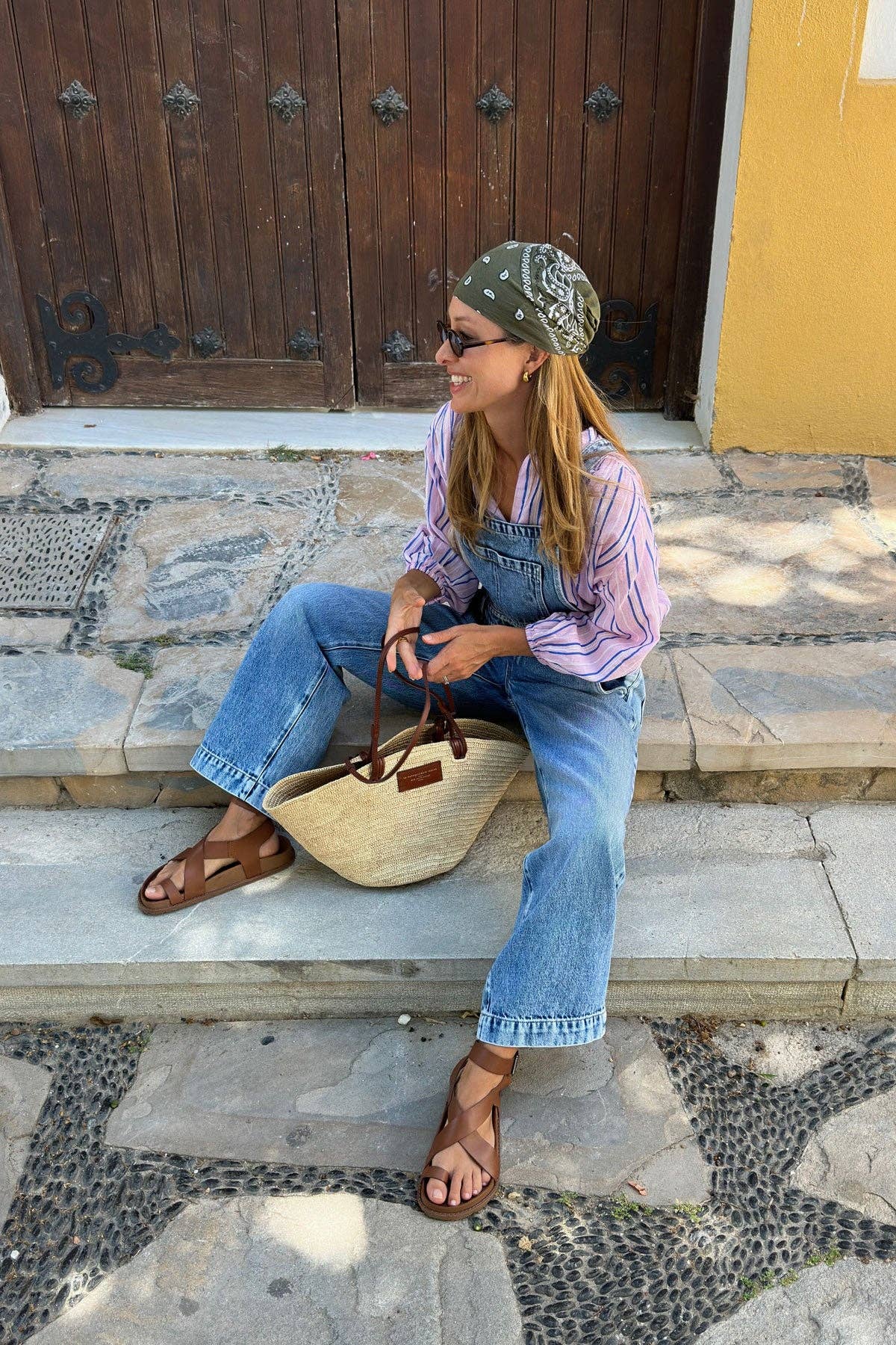 A woman sits on stone steps, smiling. She wears a green bandana, sunglasses, striped shirt, denim overalls, and Mexas EU Tampico sandals while holding a straw tote bag. A wooden door and yellow wall are behind her.
