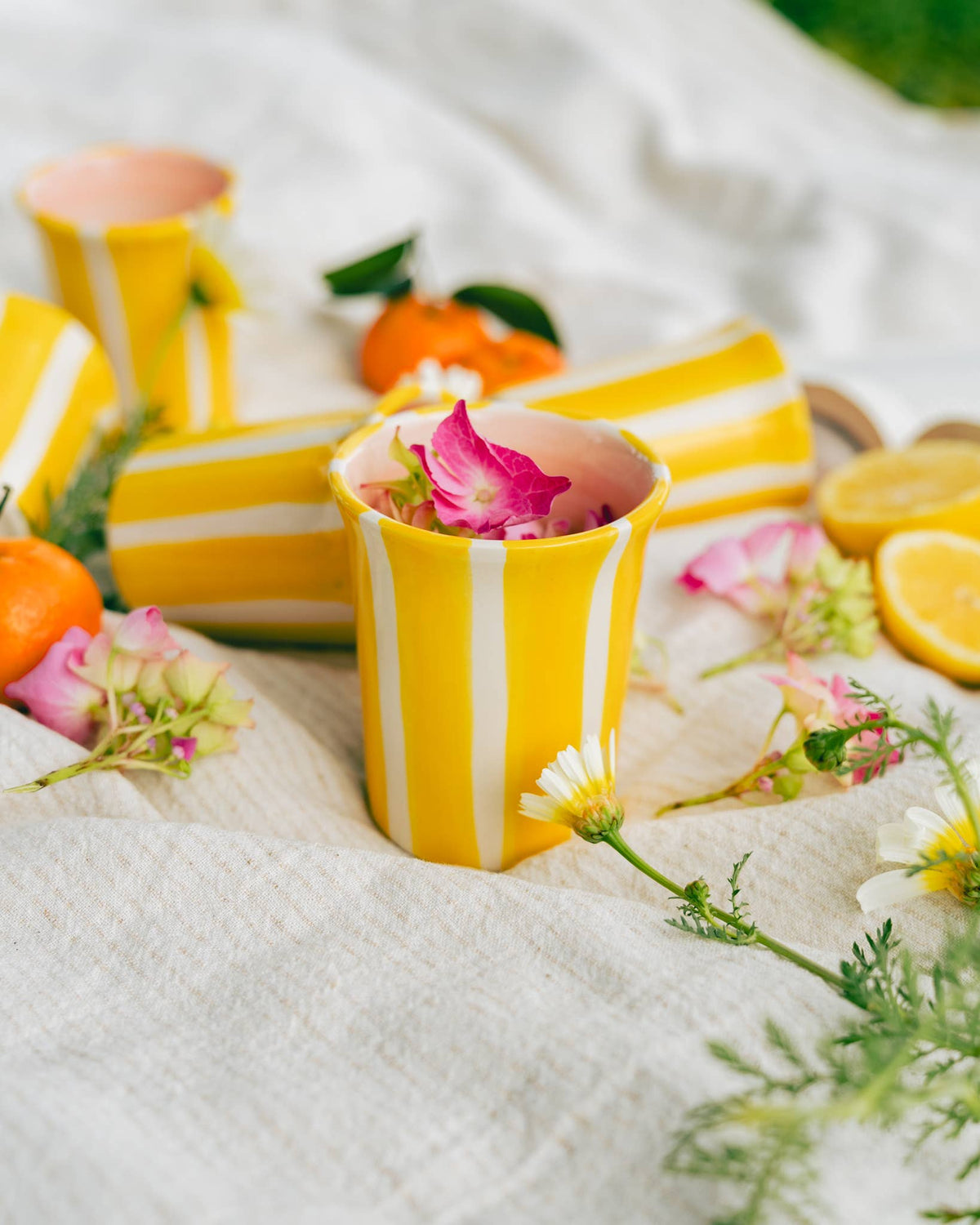 Several Valentina Handmade Spanish Ceramic Mugs – Pink & Yellow Stripes scattered on a textured fabric surface, surrounded by pink flowers, citrus fruits, and greenery.