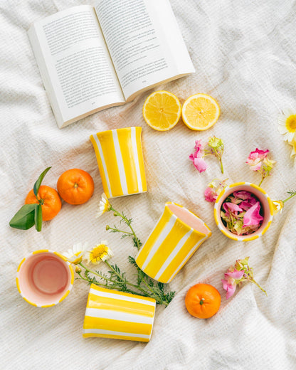 Three Valentina Handmade Spanish Ceramic Mug – Pink & Yellow Stripes mugs arranged on a textured cloth with scattered oranges, lemon slices, and flower petals, next to an open book.